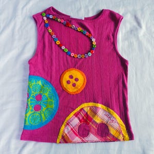 May include: A sleeveless magenta top with colorful button embellishments and a button necklace. The shirt features large, decorative button appliqués in orange, blue, and plaid patterns. The top is made of a ribbed fabric.