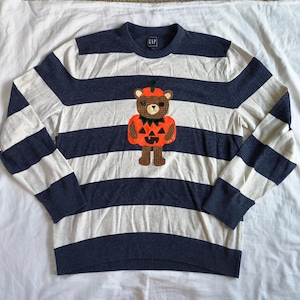 May include: A long-sleeve crewneck sweater by Gap featuring horizontal navy blue and light gray stripes. Centered on the chest is an appliqué of a brown teddy bear wearing an orange pumpkin costume with a jack-o'-lantern face and green stem.