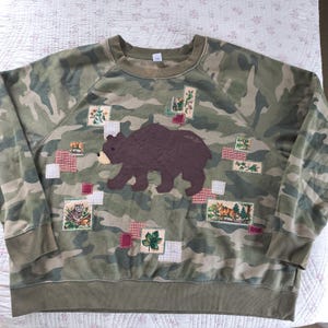 May include: A camouflage print sweatshirt featuring a large, dark brown quilted bear appliqué in the center. The garment is decorated with various small, rectangular fabric patches depicting woodland animals, leaves, and floral patterns.