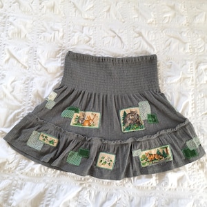 May include: A gray skirt with a ribbed waistband and a ruffled hem. The skirt features patches with images of woodland animals and floral designs. The patches are in shades of green and white.