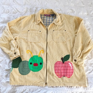 May include: A light yellow corduroy jacket with a full zipper closure. The jacket features two front pockets and is decorated with a green caterpillar and a red and white checkered apple applique. The interior lining has a blue and white plaid pattern.