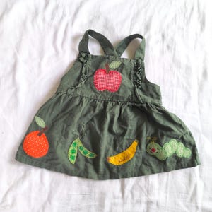 May include: An olive green sleeveless dress featuring a red and white gingham apple appliqué on the chest. The skirt displays fabric appliqués of an orange polka dot fruit, a green pea pod, a yellow banana, and a green patterned caterpillar.