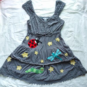 May include: A gray sleeveless dress featuring a V-neckline with eyelet lace trim, wide crochet straps, and a drawstring waist. The skirt has two tiers decorated with fabric appliqués of a red ladybug, a blue dragonfly, a green caterpillar, and yellow stars.