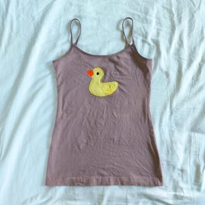 May include: A mauve-colored camisole with thin spaghetti straps. A yellow duck applique with an orange beak is centered on the front. The camisole is made of a soft, lightweight fabric, perfect for warm weather.