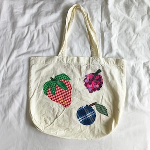 May include: Cream-colored tote bag with three appliqued fruit designs: a red and white checkered strawberry, a purple and pink raspberry, and a blue plaid plum. The bag has two cream-colored handles.