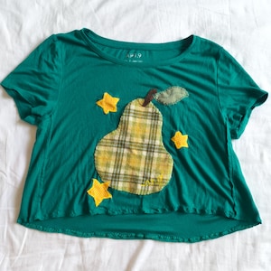 May include: A teal short-sleeved t-shirt featuring a central fabric applique of a plaid pear with a brown stem and a green leaf. The design includes three yellow star-shaped fabric appliques surrounding the pear. The shirt has a crew neckline.