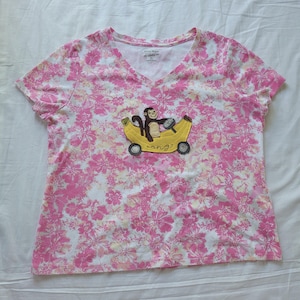 May include: A short-sleeved, V-neck t-shirt with a pink floral pattern. The shirt features an embroidered design of a monkey driving a banana car. The text "ang?" is below the banana car.