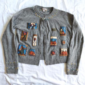 May include: A grey long-sleeve button-down cardigan featuring embroidered floral patterns around the neckline and cuffs, decorated with various rectangular fabric patches depicting scarecrows, pumpkins, and autumn-themed imagery.