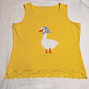 May include: A yellow tank top with a white goose applique. The goose has a blue patterned bonnet, an orange beak and feet, and the word "ang" on its body. The bottom hem features a yellow lace trim.