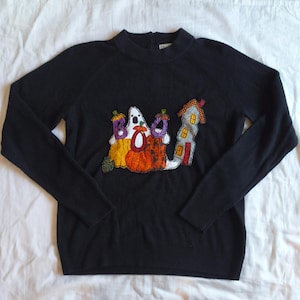 May include: A black long-sleeved jumper featuring a central appliqué design of a white ghost, orange and yellow pumpkins, and a tall house. The word BOO is spelled out with the ghost and pumpkins, with purple and orange accents on the letters.