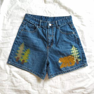 May include: High-waisted blue denim shorts featuring a button and zipper fly closure. The front panels are decorated with fabric appliques, including two green plaid pine trees on the left and a brown bear with a green plaid pine tree on the right.
