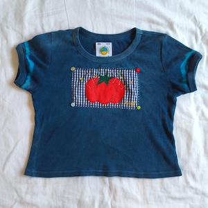 May include: A dark blue ribbed short-sleeve t-shirt featuring a rectangular patch with a black and white gingham pattern. The patch displays a red tomato graphic with green leaves and is accented by four small colorful buttons at the corners.