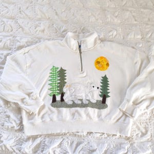 May include: White sweatshirt with a quarter-zip neckline. The front features an appliqued design of a polar bear, trees, and a yellow sun. The trees are green and the bear is white. The sweatshirt has long sleeves.