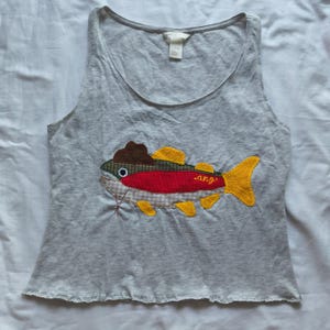May include: A light gray sleeveless tank top featuring a large, colorful fabric appliqué of a fish wearing a brown hat. The fish is constructed from various patterned fabrics in red, yellow, green, and beige, with the word ang embroidered in yellow.