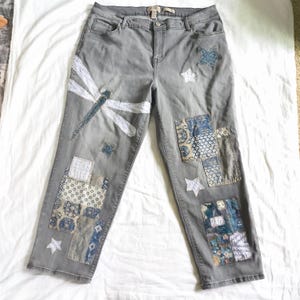 May include: Gray denim jeans featuring a large white embroidered dragonfly on the left leg, various blue and white patterned fabric patches, and several small white and blue star-shaped appliqués scattered across the front.