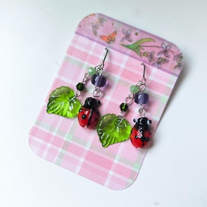 May include: A pair of ladybug earrings with green leaf charms and purple and green bead accents. The ladybugs are red with black spots and black heads. The earrings are displayed on a pink and white plaid card.