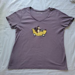 May include: A mauve V-neck t-shirt featuring an embroidered design of a monkey driving a banana car. The banana car has wheels and the word "mango" is written on the side. The shirt is made of a soft material.