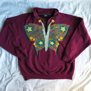 May include: A burgundy sweatshirt with a butterfly design. The butterfly is made of olive green fabric with yellow stars and colorful stitching. The sweatshirt has a zipper at the collar.