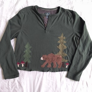 May include: A dark olive green long-sleeve henley shirt featuring a raw-edge hem. The front displays fabric appliqués of two evergreen trees, a brown bear, and three small red mushrooms. The neckline has a three-button placket with brown buttons.