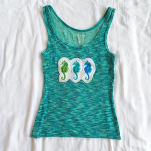 May include: A teal tank top with a scoop neck and a ribbed texture. The top features an embroidered patch of three seahorses in green, teal, and blue. The tank top is made of a soft, lightweight material.