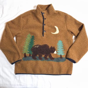 May include: Brown fleece pullover with a high collar and three buttons. The design features a bear, trees, and a crescent moon applique. The trim is navy blue, and the word "ang" is visible.