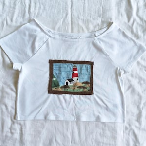 May include: A white short-sleeve t-shirt featuring a rectangular fabric appliqué on the chest. The appliqué depicts a red and white lighthouse, a small white house, and green hills against a blue sky, all enclosed within a brown border.