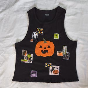 May include: A black sleeveless tank top featuring a large central orange jack-o'-lantern appliqué. The shirt is decorated with various smaller fabric patches depicting Halloween themes, including a ghost, a mummy, Frankenstein, a black cat, and candy corn.