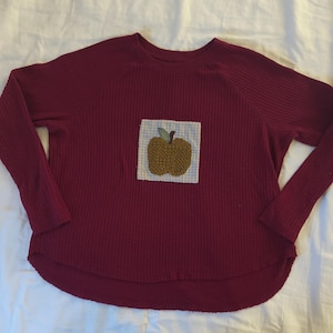 May include: A long-sleeve maroon waffle-knit shirt featuring a square patch on the chest. The patch displays a light blue and white gingham background with a textured, golden-brown apple appliqué that has a small green leaf on top.