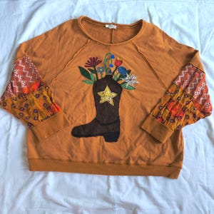 May include: A mustard-colored long-sleeve sweatshirt featuring a dark brown cowboy boot appliqué on the front. The boot is decorated with a yellow star and contains embroidered flowers. The sleeves have patchwork patterns in orange and white.