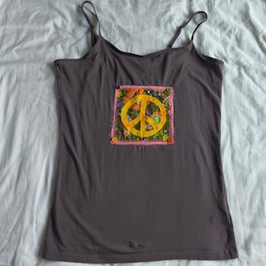 May include: A dark gray camisole top with thin straps. The top features a square patch with a yellow peace sign, surrounded by colorful flowers and a pink border. The patch is sewn onto the front of the top.