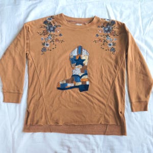 May include: A mustard-colored long-sleeve sweatshirt featuring a central patchwork cowboy boot appliqué with a dark blue star. The shoulders are decorated with embroidered floral patterns in shades of blue, white, and brown.