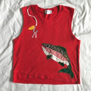 May include: Red sleeveless top with a fish applique. The fish features a patchwork design with red and white gingham, green, and gray patterns. A fishing lure and hook are appliqued near the neckline. The top has a ribbed texture.
