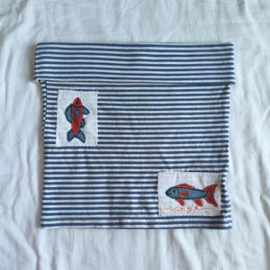 May include: A blue and white horizontally striped fabric tube with a folded top edge. Two rectangular white patches are sewn onto the fabric, each featuring an embroidered fish in red and blue. The bottom fish patch includes the word ange.