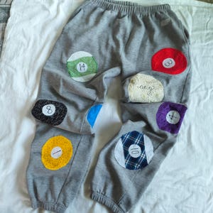 May include: Gray sweatpants with colorful patches resembling billiard balls. Patches include numbers 1, 3, 4, 8, 10, and 14, along with a patch with the word "ang". The sweatpants have an elastic waistband and cuffs.