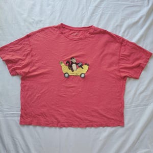 May include: Coral-colored t-shirt featuring an embroidered design of a monkey riding a banana-shaped vehicle. The monkey is brown and wearing a blue hat. The banana vehicle has yellow wheels and the word "ang" is written on the banana.