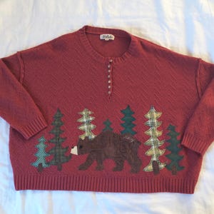 May include: A brick-red long-sleeve sweater with a textured chevron knit pattern, featuring a button-up henley neckline and a label reading Listicle. The front displays appliquéd fabric pine trees and a brown bear in the center.