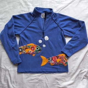 May include: Blue long-sleeved pullover with a textured pattern. The pullover features two colorful fish appliqués with floral patterns and orange tails. White bubble appliqués are near the zipper.