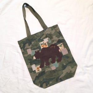 May include: Green camouflage tote bag with a brown bear applique and patches. The bag has olive green handles and the word "sang" is written on the bear. The bag is made of a durable fabric.
