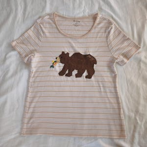 May include: Tan and white striped t-shirt with a brown bear applique holding a colorful fish. The bear has the word "ang" embroidered on its back. The shirt has a scoop neck and short sleeves.
