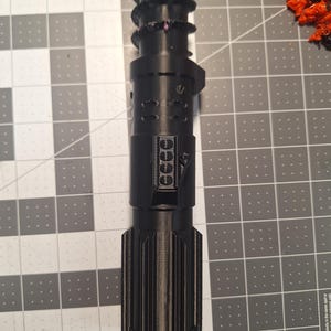 May include: A black, futuristic-looking device with a cylindrical body and textured grip. The device has multiple sections with detailed designs, including a series of small, circular features and a ribbed lower portion. The overall aesthetic suggests a sci-fi or fantasy theme.