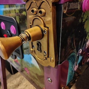 May include: A close-up of a pinball machine featuring a gold-colored, anthropomorphic door handle. The machine has a purple and black frame with a colorful Alice in Wonderland-themed graphic. Pink buttons and a control panel are visible.