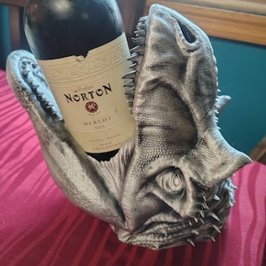 May include: A silver-colored dinosaur head wine bottle holder with a bottle of red wine. The wine bottle has a white label with black text that reads "Bodega Norton Merlot 2003 Argentina". The holder is on a red tablecloth.