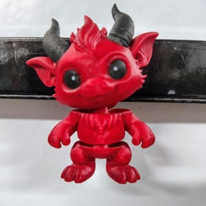 May include: A red 3D-printed figurine of a cartoon devil with large black eyes and black horns. The figurine has a detailed design, including a textured body and a small tail. The devil is posed in a standing position.