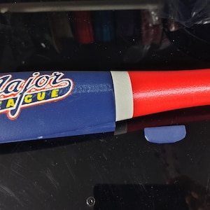 May include: A toy baseball bat featuring a blue barrel with the text Major League printed in white and yellow, a white ring, and a bright red handle. The bat has a textured surface and is positioned horizontally against a dark, reflective background.