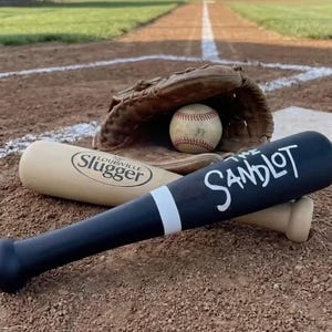 May include: A brown leather baseball glove containing a white baseball with red stitching, positioned behind a black baseball bat with the white text The Sandlot and a light tan baseball bat with the black text Louisville Slugger.
