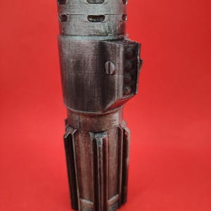 May include: A metallic silver and gray prop, possibly a lightsaber hilt, against a red background. The prop has a cylindrical design with detailed ridges, vents, and a rectangular side feature with circular details. The overall aesthetic suggests a science fiction theme.