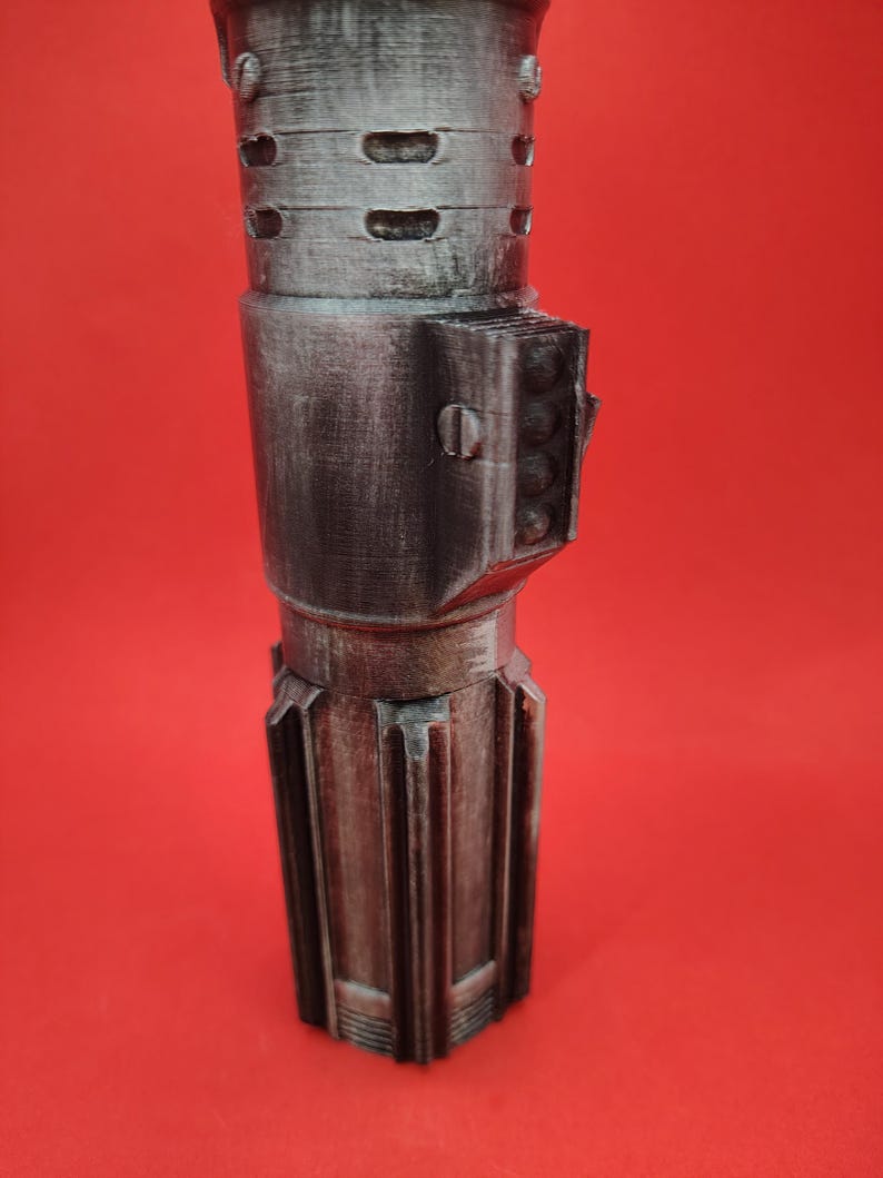 May include: A metallic silver and gray prop, possibly a lightsaber hilt, against a red background. The prop has a cylindrical design with detailed ridges, vents, and a rectangular side feature with circular details. The overall aesthetic suggests a science fiction theme.