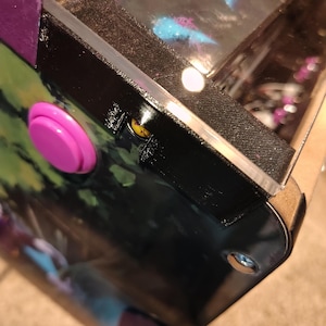May include: Close-up of a pinball machine's corner, featuring a vibrant pink button and a black, textured side panel. The machine's surface displays colorful artwork, with a purple accent on the edge. A clear plastic shield protects the playfield.