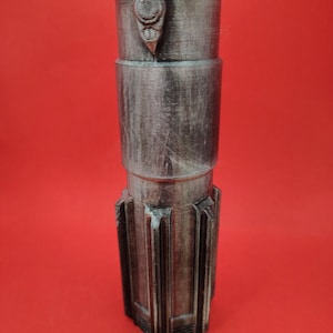 May include: A metallic gray, cylindrical object with a textured surface and a series of ridges at the base. The object has a detailed design with a button-like feature near the top. The background is a solid red.
