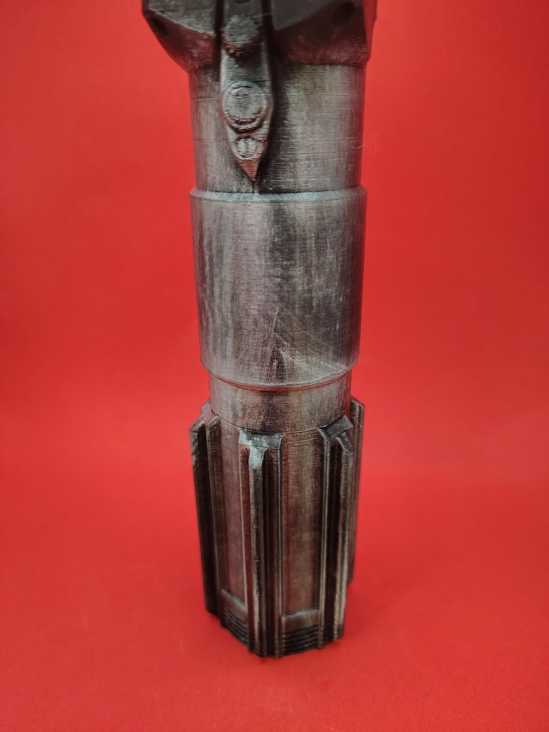 May include: A metallic gray, cylindrical object with a textured surface and a series of ridges at the base. The object has a detailed design with a button-like feature near the top. The background is a solid red.